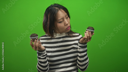 Bild auf Leinwand Woman holding two chocolate cupcakes in bare hands inspects one and bites lip in studio; indecision