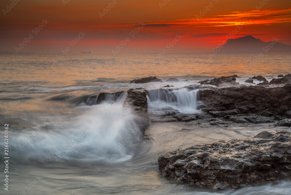 Fototapeta premium Sunbeams breaking through dark storm clouds over the sea with distant ships and rocky shore at sunset in Miono Cantabria