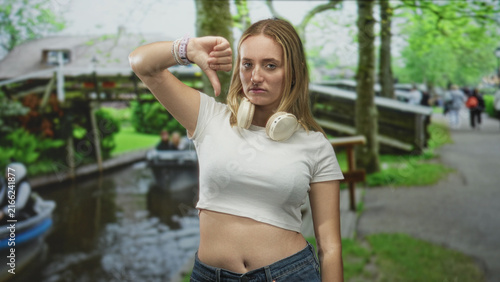 Fotografía Woman with headphones and raised arm making thumbs down gesture on street beside canal and wooden bench; disapproval defiance