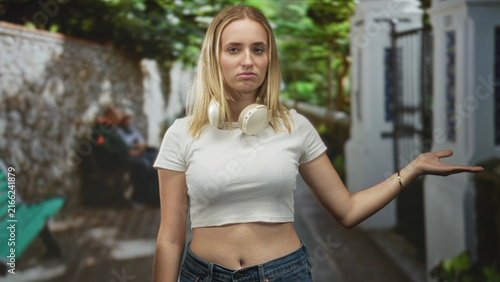 Bild auf Leinwand Woman with headphones showing palm up gesture and exposed midriff on narrow street beside white wall and green bench; indifference waiting