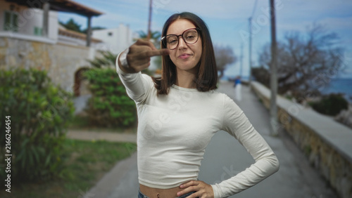 Young woman in white shirt flips middle finger to camera on a seaside street promenade, hand extended and smirking; rebellion defiance.