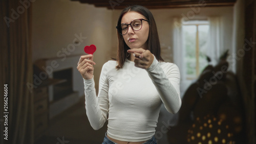 Foto Woman holds small red heart and points finger at viewer inside a building livingroom; longing relationship