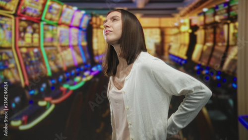 Cuadro en lienzo Young woman holding her lower back, grimacing and leaning forward beside glowing slot machines in a casino building; pain