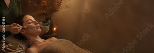 Woman receiving a relaxing facial clay mask treatment at a wellness spa. Natural skincare ritual with warm candlelight, botanical decor and copy space for beauty banner design.
