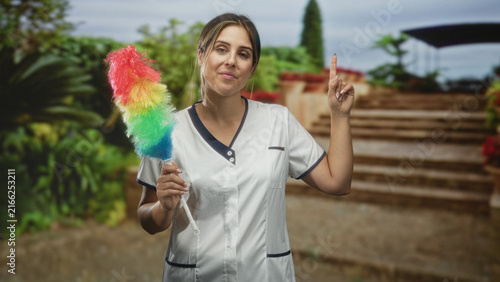 Canvastavla Woman cleaner holding rainbow feather duster and pointing finger up beside building steps in a courtyard, smiling and poised; cheerful service