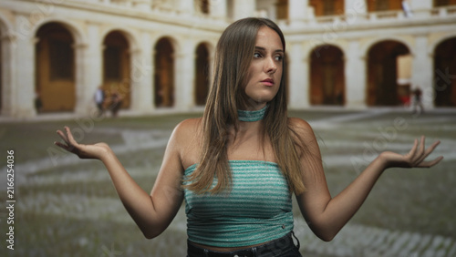 фотографія Woman shrugging with palms up and open hand gesture in building courtyard wearing teal strapless top; confusion doubt