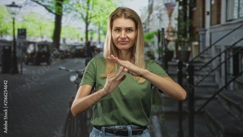 Принти на полотні Woman with hands forming a stop gesture on a street beside bicycles and stairs, wearing green t shirt and jeans; disapproval
