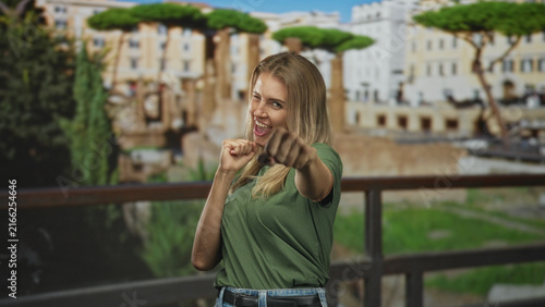 Canvas Print Blonde woman holding fists near chest on a building balcony, wearing green shirt and jeans, smiling and making a playful victory gesture; joy celebration