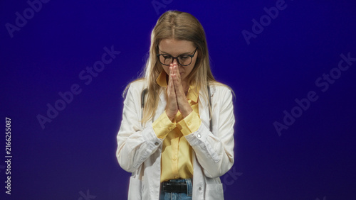 Quadro su tela Blonde woman doctor in white coat with stethoscope presses hands together in prayer gesture in a studio; hopeful reflection