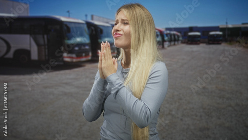 Foto Young blonde woman with hands clasped in prayer beside parked tour coaches and buses at a bus depot on a street; urgent plea hope