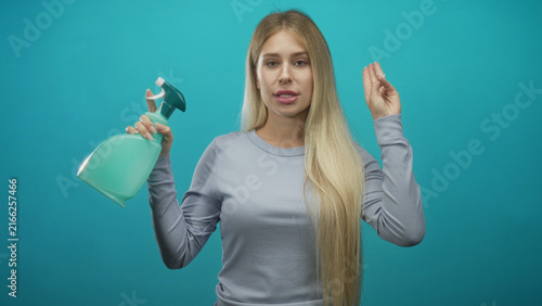 Konstfotografi Woman with long blonde hair holding teal spray bottle, fingers crossed gesture while looking at camera in studio; cleaning confidence