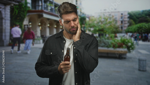 Foto Man with beard holding beer bottle touches cheek for toothache on street; discomfort hesitation uncertainty