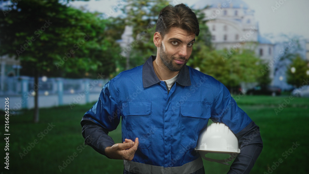 Fototapeta premium Man construction worker holding hardhat under arm and points finger to chest at building; confidence duty.