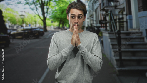Принти на полотні Man with hands pressed together in a pleading prayer gesture on a street near townhouse steps and parked cars; apology