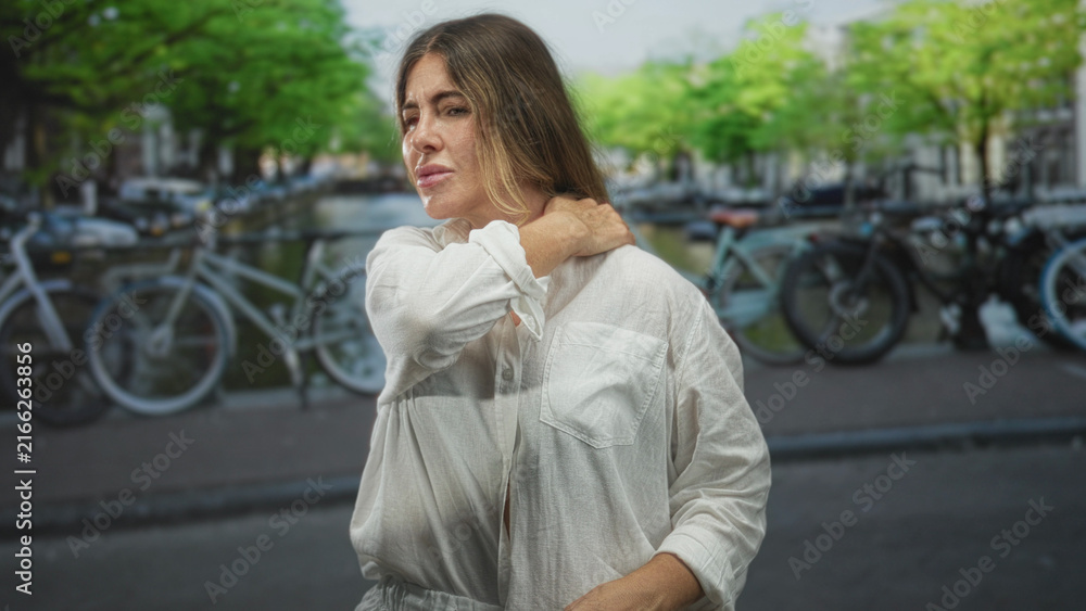 Fototapeta premium Woman with hand on neck rubbing neck in a street by a canal lined with parked bicycles, eyes closed and wincing; pain tension vulnerability.