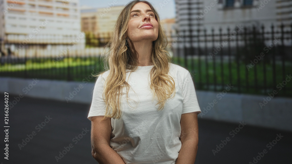 Fototapeta premium Woman in white t shirt gazes upward with closed eyes and slight smile, forearms exposed, standing on street by iron fence and city buildings; serene reflection.