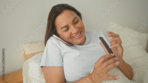 Fotografía Woman filing nails with emery board while holding smartphone to ear on bedroom bed; relaxed self care