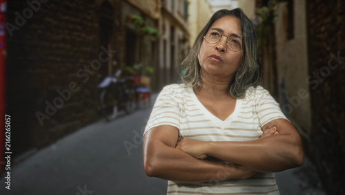 фотографія Hispanic middleaged woman with glasses standing on a narrow street, arms crossed and folded across chest, stern expression and direct gaze; stubbornness resolve