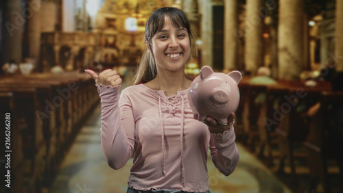 Canvastavla Young woman in pink sweatshirt holding ceramic piggybank and pointing thumb to her chest while standing in a church aisle; financial confidence