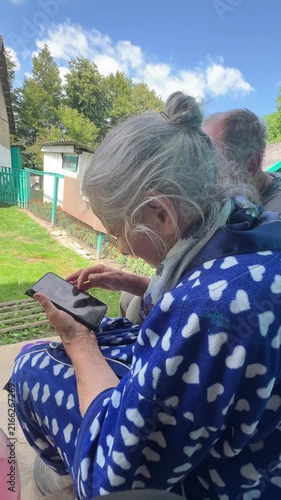 Senior woman in heart patterned robe using smartphone outdoors in rural backyard, grey hair in bun, technology for elderly, side view
