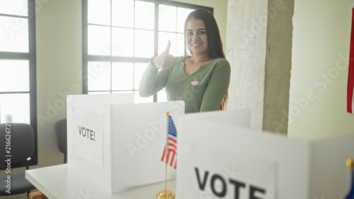 Fotografía Young hispanic woman gives thumbs up at a voting booth with i voted sticker and small us flag in building; civic pride