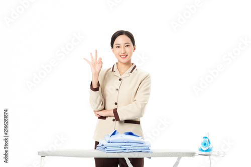 Foto Smiling housekeeper with folded clothes and iron making okay gesture
