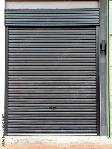 Black metal roller shutter door closed at a shop entrance. Industrial security gate with horizontal slats and a lock. Modern storefront exterior with locked garage door and concrete sidewalk detail.