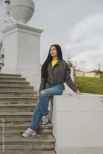 A young Asian woman with long black hair sits on a staircase. She wears a gray jacket, yellow shirt, and ripped jeans, with sneakers on her feet.