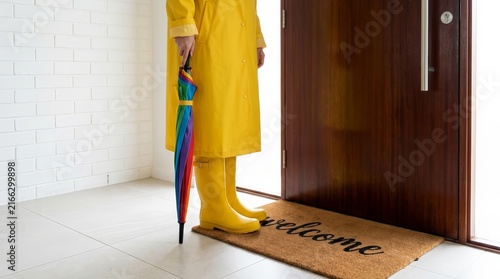Woman standing at a home entrance in a yellow raincoat with a rainbow umbrella and gumboots.