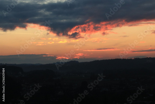 Spektakulärer Sonnenuntergang am Horizont über einer dunklen Hügellandschaft. Der Himmel ist in dramatische orangefarbene und rote Töne getaucht, während dunkle Wolken das Lichtspiel intensivieren.
