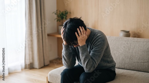 Stressed young man sitting on sofa holding head in hands, suffering from headache or migraine pain, mental health crisis, depression and anxiety disorder symptoms