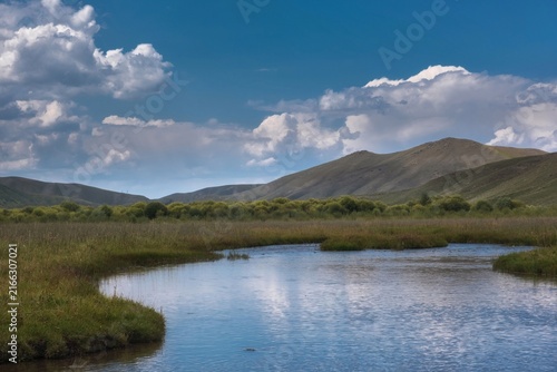 A quiet river stream flowing through a green meadow with rolling hills under a blue sky filled with white clouds. Suitable for country lifestyle, eco tourism, and natural background designs.
