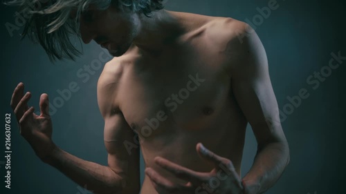 feel your strength Shirtless young man with long hair posing with raised hands in low-key lighting against a dark studio background, showcasing a muscular upper body and contemplative expression