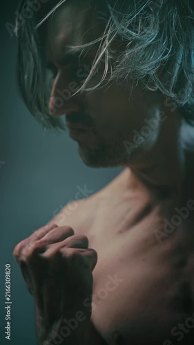 to fight with oneself Close-up artistic portrait of a shirtless young man with long tousled hair, expressive profile and hand gestures in low-key lighting against a soft gray-blue background