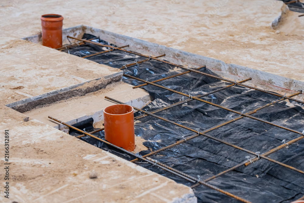 Naklejka premium Subgrade foundation construction showing a steel rebar grid, waterproof vapor barrier sheeting, and orange PVC utility pipes for plumbing