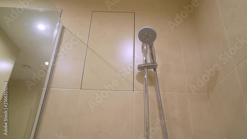 Modern chrome shower head dispensing water stream against beige marble tiled wall. Clean minimalist bathroom interior with soft lighting reflective surfaces