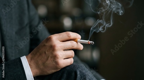 Elegant hand in suit sleeve holding burning cigarette with rising smoke trail in moody warm lighting.
