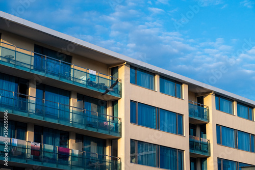 A modern residential apartment building with glass balconies bathed in warm sunlight against a partly cloudy blue sky