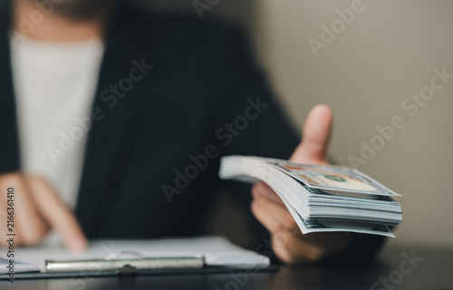 A professional person in formal attire holds a stack of currency bills and financial documents, indicating financial transactions or business activities in an office setting.