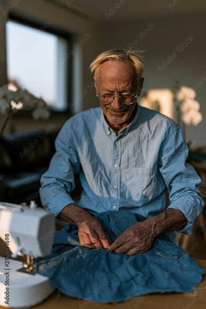 Obraz premium Elderly man hand sewing by a domestic sewing machine at home for repair and slow craft hobby, warm lifestyle background for diy or wellness content