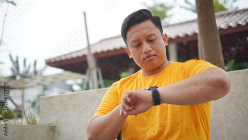 Young Asian man in a yellow shirt looking at his black smartwatch on the wrist, checking time or fitness notification outdoors. Wearable technology and healthy lifestyle concept in a tropical setting.