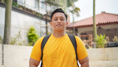 Portrait of a confident young Asian man in a yellow shirt wearing a backpack, standing outdoors looking at the camera. Casual lifestyle concept of a student or traveler in a tropical neighborhood.