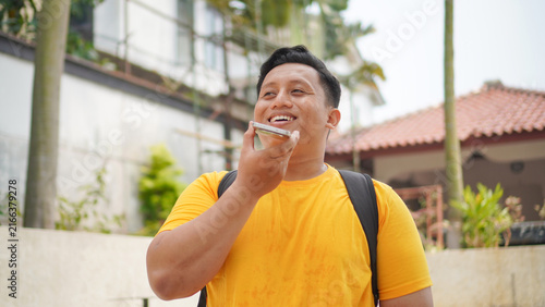 Person in a yellow shirt using an AI voice assistant on a smartphone, speaking a voice command to the glowing orb on screen showing 