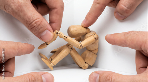 Wooden mannequin crouching in a corner surrounded by pointing fingers, conceptual image representing bullying, social pressure, harassment, loneliness, and judgment.