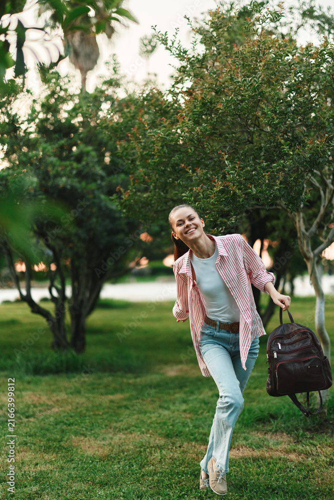 Fototapeta premium Smiling student woman running in a park with a backpack, casual daytime scene conveying study life, energy, and outdoor activity. The image captures youthful ambition and everyday campus lifestyle
