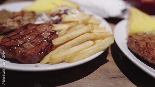 Grilled pork steak served on a white plate with crispy golden french fries, savory gravy sauce, and a fresh side salad. Close-up view of a delicious restaurant meal under warm ambient lighting.