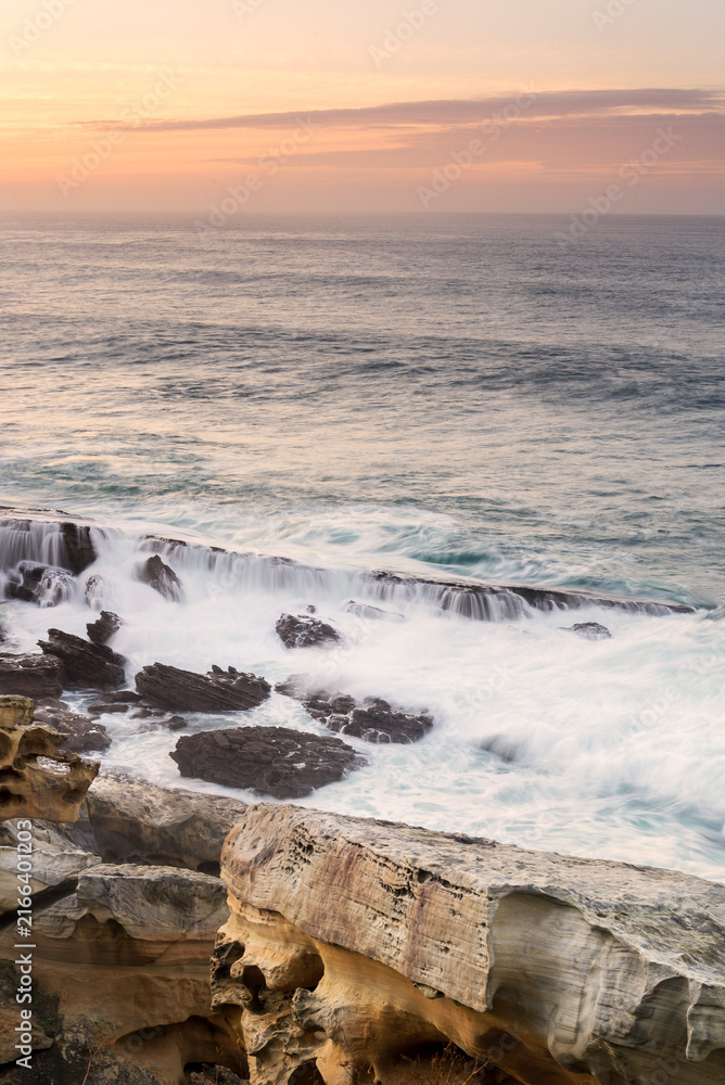 Fototapeta premium Sandstone rock formations with tafoni erosion on the coast of Jaizkibel during a dramatic sunset with long exposure ocean waves flowing over rocks in Jaizkibel, Basque Country, Spain