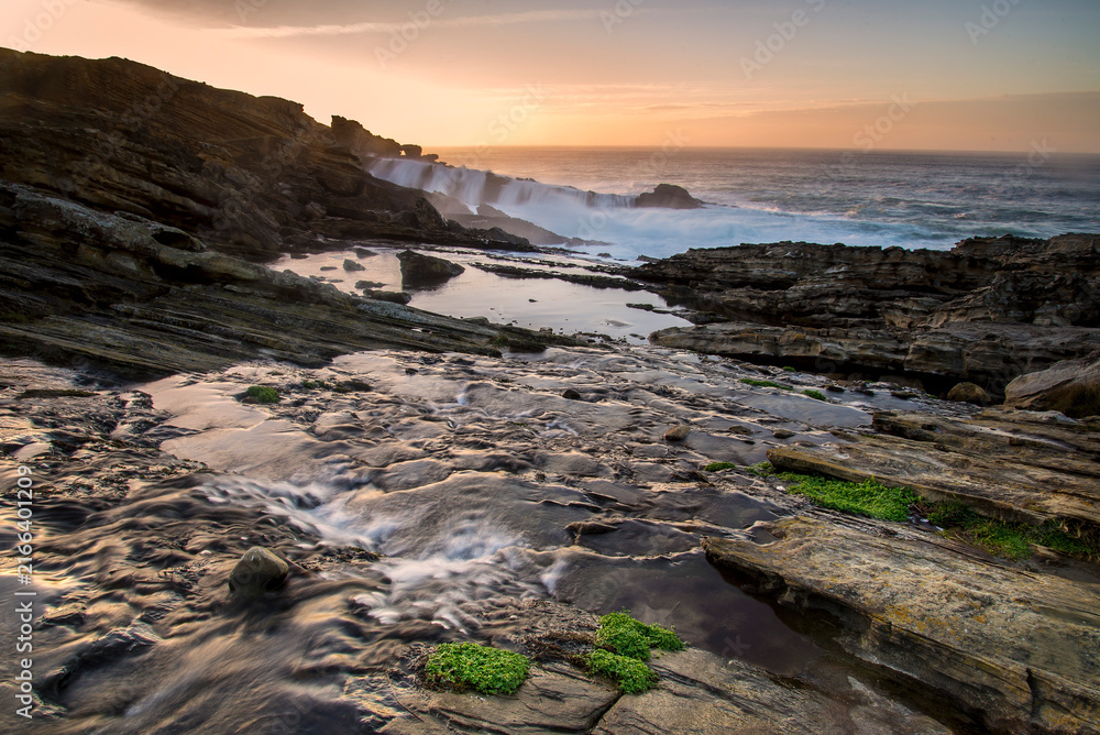 Fototapeta premium Sandstone rock formations with tafoni erosion on the coast of Jaizkibel during a dramatic sunset with long exposure ocean waves flowing over rocks in Jaizkibel, Basque Country, Spain