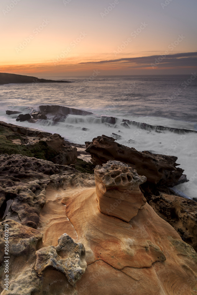 Fototapeta premium Sandstone rock formations with tafoni erosion on the coast of Jaizkibel during a dramatic sunset with long exposure ocean waves flowing over rocks in Jaizkibel, Basque Country, Spain