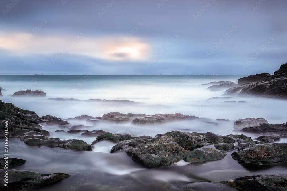 Fototapeta premium Dramatic pink sunset sky with long exposure ocean waves washing over a dark pebble beach and coastal hills in Ogeia Beach